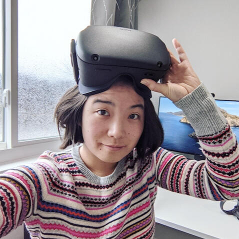 Photograph of an Asian woman wearing a striped sweater holding VR goggles over her head